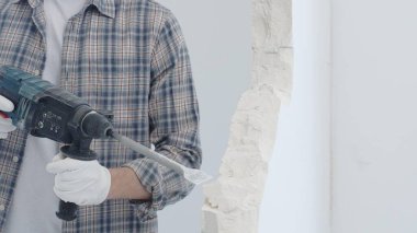 İnşaat işçisi, beyaz duvarı döner çekiçli matkapla yıkıyor, gri kareli gömlek ve koruyucu beyaz eldivenler giyiyor, yenileme projesi sırasında toz üretiyor, yakın plan görüntüsü var..
