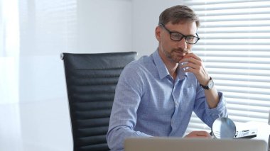 Yetişkin erkek muhasebeci vergi belgelerini incelerken düşünüyor, üst üste dizilmiş dosyaların yanında hesap makinesi kullanıyor, açık mavi gömlekli siyah ofis koltuğunda oturuyor. Denetim ve vergiler.