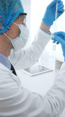 Laboratuvar şapkası, yüz maskesi, Google ve mavi eldiven takan erkek tıp bilimci modern bir laboratuvar ortamında bir deney tüpünün içine dikkatlice sıvı bırakıyor. Tıp, sağlık hizmeti kavramı.