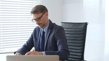 Kıdemli işadamı takım elbise, kuyruk ve siyah gözlük takarak masasındaki parlak, modern bir ofiste profesyonelliği gösteren bir belgeye konsantre oluyor. İş adamları konsepti.