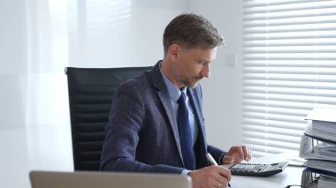 Takım elbiseli ve kravatlı olgun bir iş adamı ofis masasında oturuyor ve hesap makinesi kullanarak evrakları imzalıyor. İş dünyasında denetim ve vergi teması