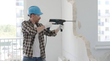 İnşaat işçisi, koruyucu teçhizatlı bir çekiç matkabıyla duvarı yıkıyor ve zemin yüzeyinde inşaat enkazı oluşturuyor. Yenileme konsepti.