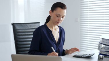 Masada hesap makinesiyle oturan ve not alan, modern ofis ortamında finansal verileri ve evrak işlerini yöneten odaklı bir iş kadını. İş dünyasında denetim ve vergi teması