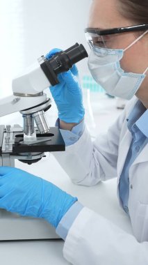 Mavi koruyucu giysili kadın bilim adamı laboratuvar araştırma ortamında test örneklerini mikroskop altında analiz ediyor. Tıp, sağlık ve bilim konsepti.