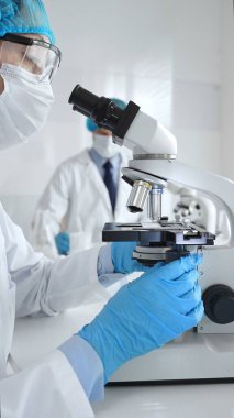 Maske takan ve mavi güvenlik donanımı takan kadın bir bilim adamı araştırma için mikroskop kullanırken erkek bir meslektaşı arka planda çalışıyor. Tıp, sağlık ve bilim konsepti.