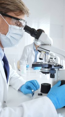 Mikroskop kullanarak maske takan ve eldiven giyen erkek bilim adamlarına yakın çekim, arka planda çalışan meslektaşlarıyla modern laboratuvarda araştırma yapıyorlar. Tıp, sağlık ve bilim konsepti