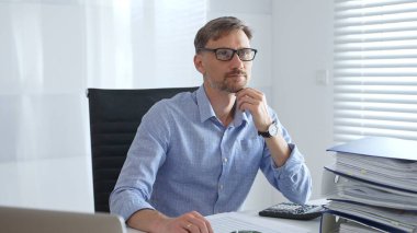 Ciddi bir erkek muhasebeci vergi belgelerini incelerken, dizilmiş dosyaların yanında hesap makinesi kullanarak, açık mavi gömlekli, siyah ofis koltuğunda oturarak düşünüyor. Ticarette denetim ve vergiler.