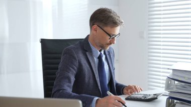 Takım elbiseli, kravatlı ve gözlüklü olgun girişimci masada oturuyor, notlar yazıyor ve modern, parlak ofiste hesap makinesi kullanıyor. Ticarette denetim ve vergiler.