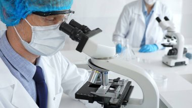 Koruyucu maske, şapka ve eldiven takan kıdemli erkek bilim adamı modern bir laboratuvarda mikroskop altında örnekleri inceliyor. Tıp, sağlık ve bilim konsepti.