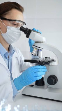 Maske takan ve mavi eldiven giyen kadın bilim adamı mikroskop kullanarak arka planda çalışan bir meslektaşla modern laboratuvarda bilimsel araştırmalar yapıyor. Tıp, sağlık ve bilim konsepti.