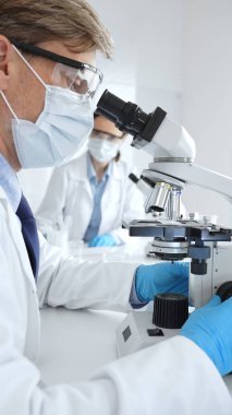 Mikroskop kullanarak maske takan ve eldiven giyen erkek bilim adamlarına yakın çekim, arka planda çalışan meslektaşlarıyla modern laboratuvarda araştırma yapıyorlar. Tıp, sağlık ve bilim konsepti