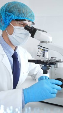 Koruyucu maske, şapka ve eldiven takan kıdemli erkek bilim adamı modern bir laboratuvarda mikroskop altında örnekleri inceliyor. Tıp, sağlık ve bilim konsepti.