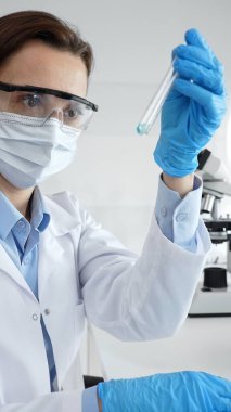 Maske takan, koruyucu gözlük takan ve eldiven takan kadın bilim adamı modern laboratuardaki tıbbi araştırma sırasında mavi sıvıyı dikkatlice inceliyor. Tıp, sağlık ve bilim konsepti