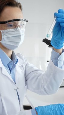 Maske takan, koruyucu gözlük takan ve eldiven takan kadın bilim adamı modern laboratuardaki tıbbi araştırma sırasında mavi sıvıyı dikkatlice inceliyor. Tıp, sağlık ve bilim konsepti