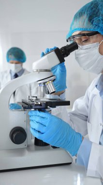 Bilinmeyen bilim adamı mavi güvenlik donanımı takıyor. Araştırma için mikroskop kullanıyor. Erkek bir meslektaşı arka planda çalışıyor. Tıp, sağlık ve bilim konsepti.