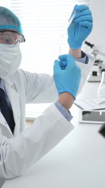 Laboratuvar şapkası, yüz maskesi, Google ve mavi eldiven takan erkek tıp bilimci modern bir laboratuvar ortamında bir deney tüpünün içine dikkatlice sıvı bırakıyor. Tıp, sağlık hizmeti kavramı.