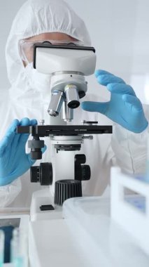 Koruyucu ekipman takan bilim araştırmacısı mikroskobu ayarlıyor, laboratuvar örneklerini steril araştırma ortamında hassasiyetle analiz ediyor. Tıp, sağlık ve bilim konsepti.