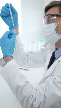 Laboratuvar önlüklü, koruyucu gözlüklü ve mavi eldivenli maskeli erkek bilim adamı modern laboratuardaki test tüplerine mikroskop ve test tüpleri rafı koyuyor. Tıp, sağlık ve bilim konsepti.