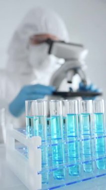 Laboratuvarda koruyucu dişli mikroskop lensleri takan bir bilimsel araştırmacının karşısında mavi sıvıyla test tüplerini kapatın. Tıp, sağlık ve bilim konsepti.