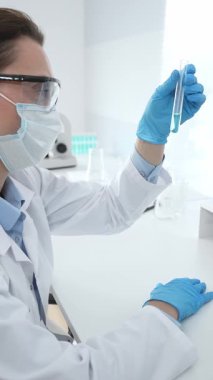 Mavi koruyucu eldivenli maskeli kadın araştırmacı test tüpündeki temiz sıvıyı inceliyor, mikroskop ve laboratuvar ekipmanlarının yanında oturuyor. Tıp, sağlık ve bilim konsepti.