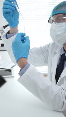 Laboratuvar önlüklü, koruyucu gözlüklü ve mavi eldivenli maskeli erkek bilim adamı modern laboratuardaki test tüplerine mikroskop ve test tüpleri rafı koyuyor. Tıp, sağlık ve bilim konsepti.