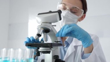 Koruyucu ekipman takan kadın bilim adamı mikroskobu kontrol ediyor, modern laboratuvar araştırma ortamında mavi test tüplerini analiz ediyor. Tıp, sağlık ve bilim konsepti.