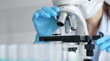 Mikroskobu ayarlayan eldivenli bilim adamlarına yakın çekim yapmak, laboratuvarda araştırma yapmak ve arka planda mavi sıvı içeren test tüpleri kullanmak. Tıp, sağlık ve bilim konsepti.