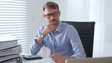 Ciddi bir erkek muhasebeci vergi belgelerini incelerken, dizilmiş dosyaların yanında hesap makinesi kullanarak, açık mavi gömlekli, siyah ofis koltuğunda oturarak düşünüyor. Ticarette denetim ve vergiler.