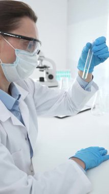 Mavi koruyucu eldivenli maskeli kadın araştırmacı test tüpündeki temiz sıvıyı inceliyor, mikroskop ve laboratuvar ekipmanlarının yanında oturuyor. Tıp, sağlık ve bilim konsepti.