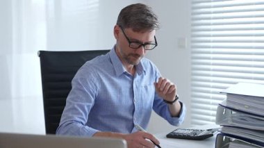 Mavi gömlekli ve gözlüklü profesyonel bir muhasebeci ofis ortamındaki mali raporları incelerken faturaları hesaplıyor ve düşünüyor. İş dünyasında denetim ve vergi teması.