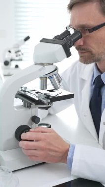 Beyaz önlük ve gözlük giyen erkek bilim adamı mikroskop kullanıyor ve modern bir laboratuvarda bilimsel araştırma yapıyor. Tıp, sağlık ve bilim konsepti.