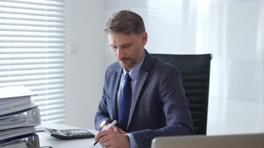 Takım elbiseli ve kravatlı bir işadamı modern ofiste mali belgelerle uğraşırken masada oturuyor ve hesap makinesi kullanıyor. İş dünyasında denetim ve vergi teması