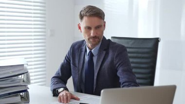 Profesyonel iş adamı dizüstü bilgisayarla uğraşırken yakın çevresindeki organize klasörlerle şık çalışma sahalarında konumlanıyor ve kurumsal ithafı vurguluyor. İş dünyasında denetim ve vergi teması