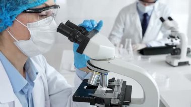 Maske takan ve mavi güvenlik donanımı takan kadın bir bilim adamı araştırma için mikroskop kullanırken erkek bir meslektaşı arka planda çalışıyor. Tıp, sağlık ve bilim konsepti.