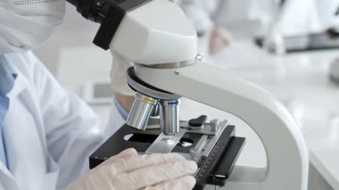 Beyaz koruyucu eldivenler giyen bilim teknisyeni laboratuvar mikroskobunu dikkatlice ayarlıyor ve titiz bir araştırma tekniği sergiliyor. Tıp, sağlık ve bilim konsepti