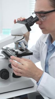 Laboratuvar önlüğü ve koruyucu gözlüklü kadın bilim adamı modern bir laboratuvarda mikroskop altında test örnekleri inceliyor. Tıp, sağlık ve bilim konsepti
