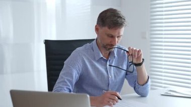 Mavi parlak gömlekli, gözlüklü bir işadamı, parlak ve modern bir ofiste çalışırken, profesyonelliğini ve verimliliğini gösteriyor. İş adamları konsepti.