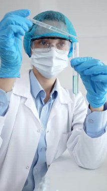 Maske takan kadın bilim adamı, mavi başlık ve eldivenler laboratuardaki pipeti kullanarak test tüpüne sıvı ekliyor. Tıp ve bilim konsepti.