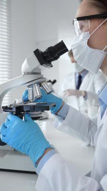 Kadın araştırmacı mikroskop ile örnek analiz ediyor, koruyucu teçhizat takılı, steril araştırma ortamında gelişmiş laboratuvar ekipmanlarının yanında çalışan bir meslektaşı. Tıp, sağlık ve bilim konsepti