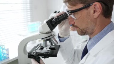 Koruyucu gözlük takan bilim adamı mikroskop altında araştırma ve analiz yapıyor. Arka planda test tüpleri ile modern bir laboratuvarda. Tıp, sağlık ve bilim konsepti