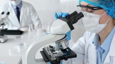 Maske takan ve mavi güvenlik donanımı takan kadın bir bilim adamı araştırma için mikroskop kullanırken erkek bir meslektaşı arka planda çalışıyor. Tıp, sağlık ve bilim konsepti.