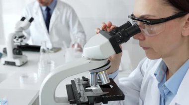 Koruyucu gözlük takan kadın bilim adamı mikroskop kullanarak modern laboratuvarda deney tüpleri ve arka planda erkek bilim adamı ile araştırma yapıyor. Tıp, sağlık ve bilim konsepti