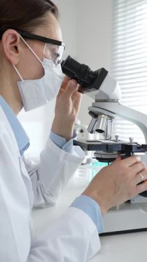 Koruyucu gözlük takan kadın bilim adamı mikroskop kullanarak modern laboratuvarda deney tüpleri ve arka planda erkek bilim adamı ile araştırma yapıyor. Tıp, sağlık ve bilim konsepti