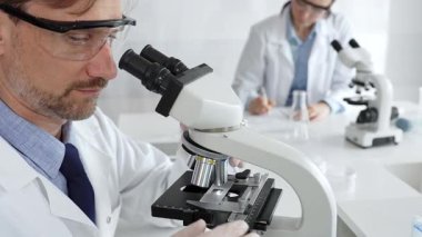 Koruyucu gözlük takan bilim adamı mikroskop altında araştırma ve analiz yapıyor. Arka planda test tüpleri ile modern bir laboratuvarda. Tıp, sağlık ve bilim konsepti