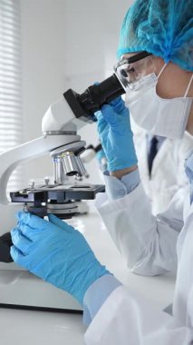 Maske takan ve mavi güvenlik donanımı takan kadın bir bilim adamı araştırma için mikroskop kullanırken erkek bir meslektaşı arka planda çalışıyor. Tıp, sağlık ve bilim konsepti.