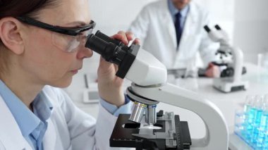 Koruyucu gözlük takan kadın bilim adamı mikroskop kullanarak modern laboratuvarda deney tüpleri ve arka planda erkek bilim adamı ile araştırma yapıyor. Tıp, sağlık ve bilim konsepti