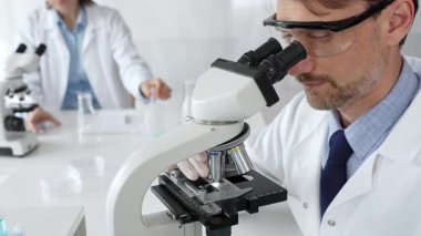 Koruyucu gözlük takan bilim adamı mikroskop altında araştırma ve analiz yapıyor. Arka planda test tüpleri ile modern bir laboratuvarda. Tıp, sağlık ve bilim konsepti