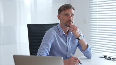 Mavi gösterişli gömlekler giyen işadamı, parlak ve modern bir ofiste masa başında çalışırken düşünüyor ve konsantre oluyor. Profesyonelliğini ve verimliliğini gösteriyor. İş adamları konsepti.