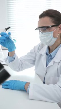 Mavi koruyucu eldivenli maskeli kadın araştırmacı test tüpündeki temiz sıvıyı inceliyor, mikroskop ve laboratuvar ekipmanlarının yanında oturuyor. Tıp, sağlık ve bilim konsepti.