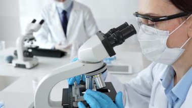 Maskeli profesyonel kadın bilim adamı mikroskop ile numune inceleyerek steril laboratuvar ortamında meslektaşlarıyla birlikte araştırma yapıyor. Tıp, sağlık ve bilim konsepti.